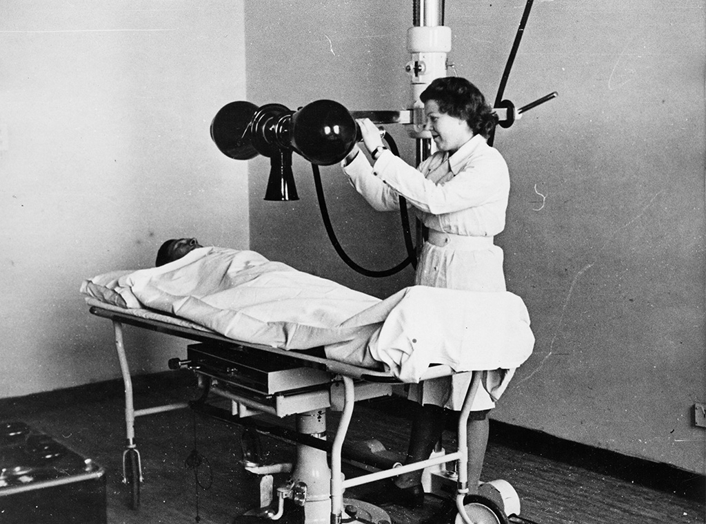 A black and white image showing a woman in a white coat standing over a patient lying down, holding an old X-ray scanner.