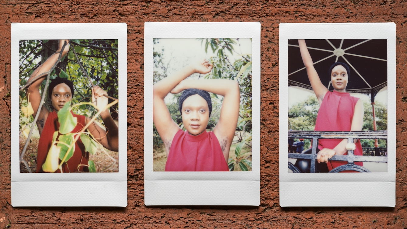 Photograph of three Instax Mini instant film prints in a line, resting on a textured brick surface. All three prints show the same woman. The print on the left shows her nestled within the branches and leaves of tree with her right arm raised up up to show her armpit hair. The print in the centre shows her from the chest up with both her arms raised over her head revealing her armpit hair. Behind her is the green leaves of shrubs and trees. The print on the right shows the same woman standing outside under a large bandstand structure. Her right arm is raised up into the air to show her armpit hair.