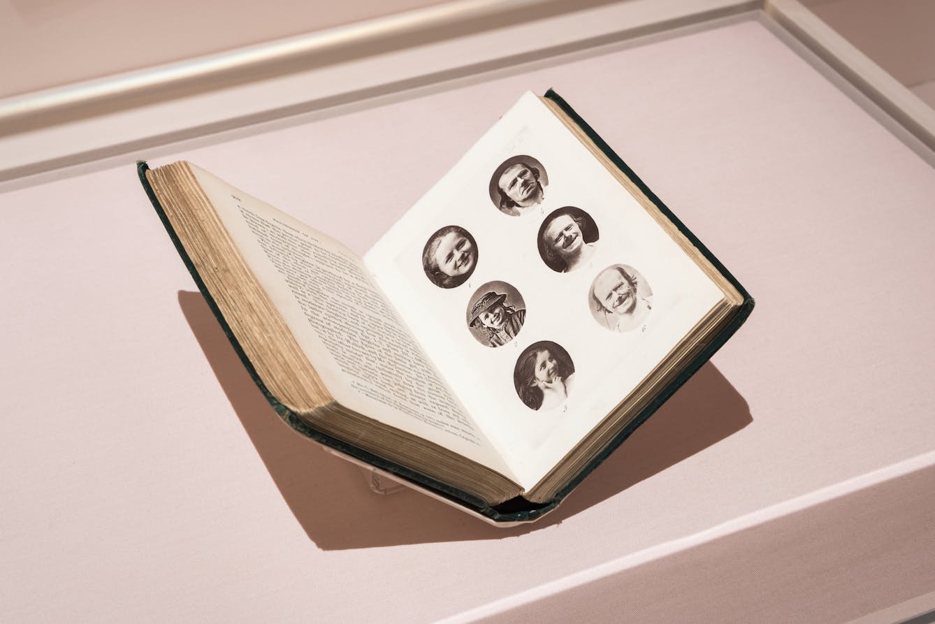 Photograph of a glass exhibition display case showing an open printed book, held in a book cradle. The open double page spread shows text on the left page and 6 circular portraits of people's expressions on the right.