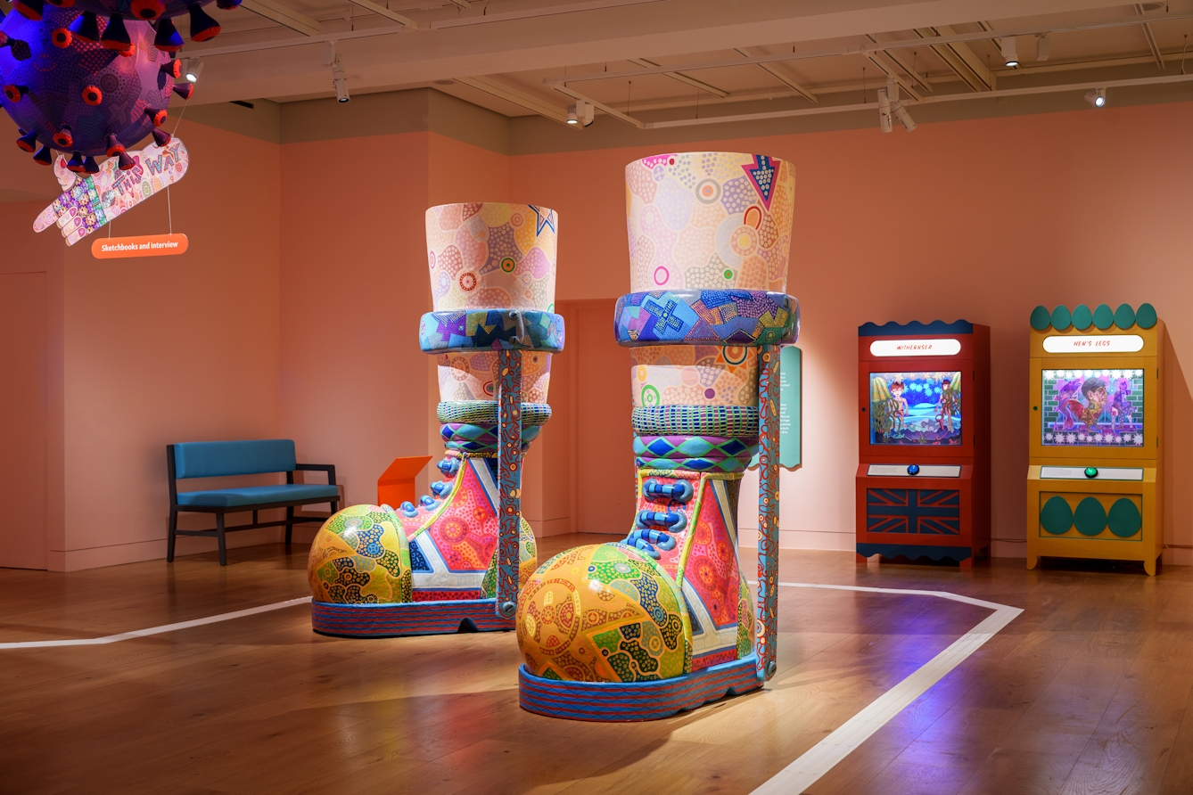 Photograph of a colourful exhibition space with  a large sculpture of a pair of boots with callipers. The sculpture is covered in colourful patterns. Behind the sculpture are 2 arcade style booths showing a multi-layered dioramas.