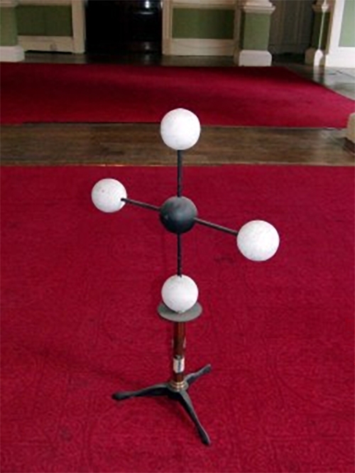 A molecular model in the shape of a cross, with a black ball at the centre, connected to four white balls.