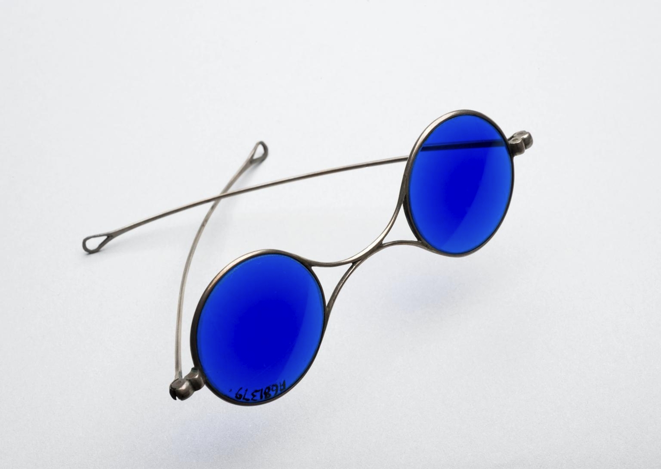 A photograph of a pair of sunglasses against a white background. The frames are made from a silver-coloured metal. The lenses are round and bright blue.