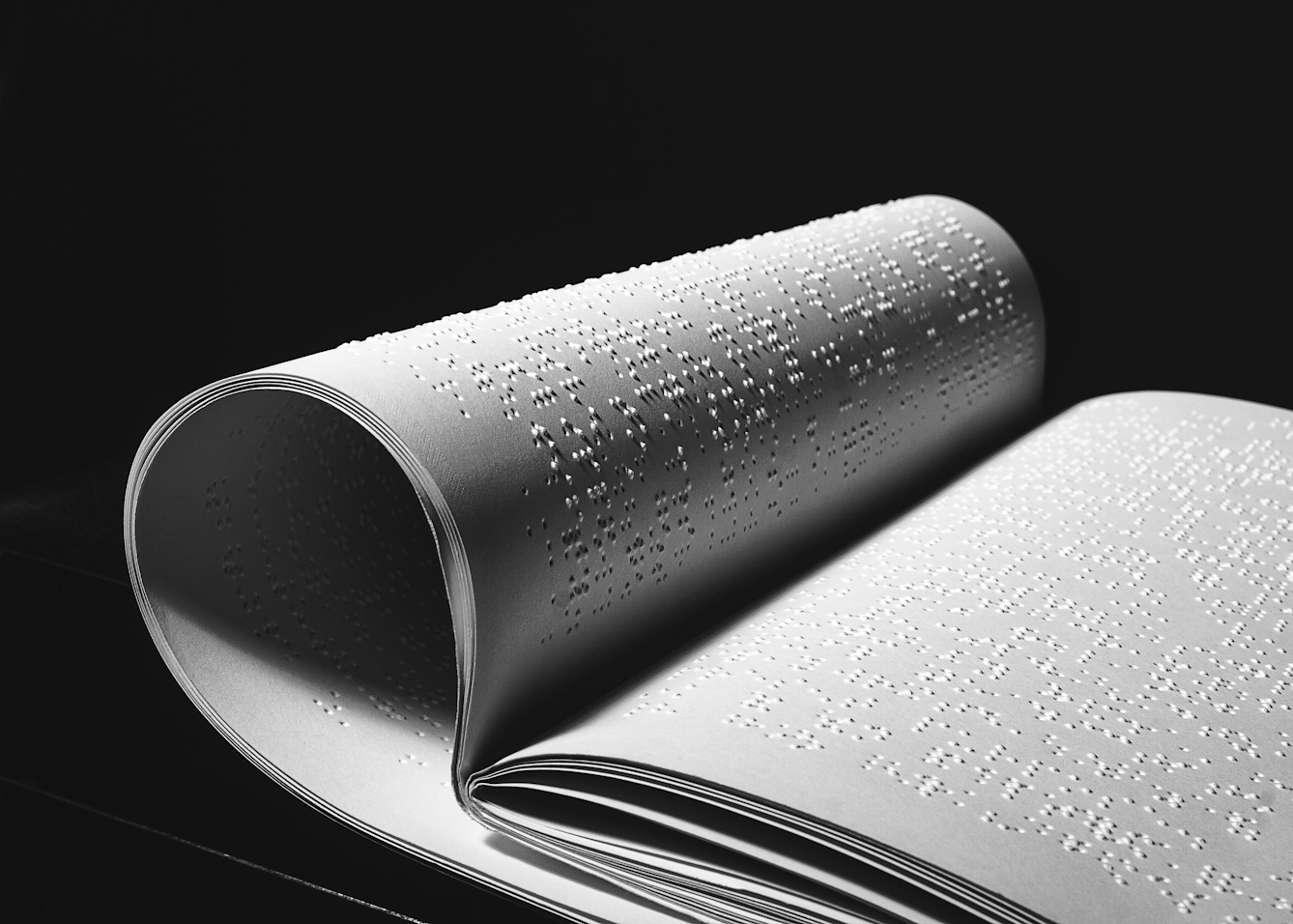 Black and white photograph of a Braille book, printed onto white sheets of paper. The sheets are bound into a book form with one half of the book curled around and tuck under the other. The sheets are lit with hard direct light which reveals the texture and form of the Braille. The background surrounding the book is plunged into darkness.