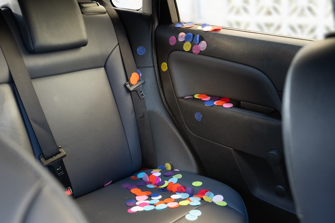 A photograph of the rear seat of a car. In the car there are multi-coloured paper circles cascading down from the window and door and pooled on the seat.