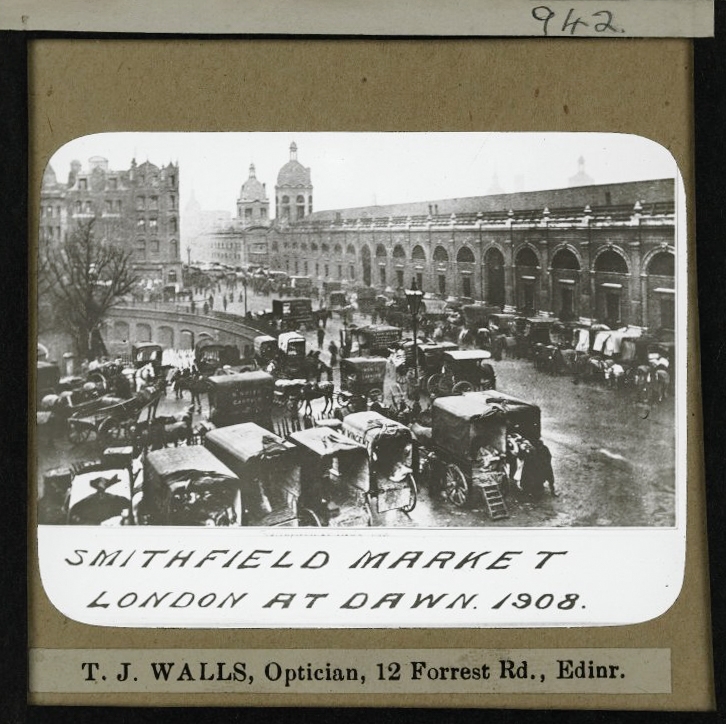Photograph of a glass slide with a handwritten caption. The slide itself shows a black and white photograph of a brick market building with many horses and carriages in the foreground.
