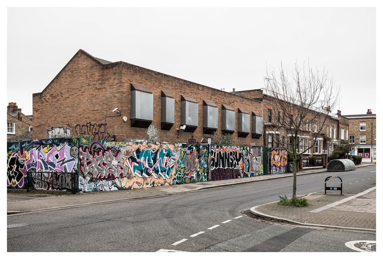 Photograph of a South London street scene in Brixton, showing a building which has all its windows boarded up with metal shutters. The building is surrounded by wooden hoarding which is covered in colourful graffiti.