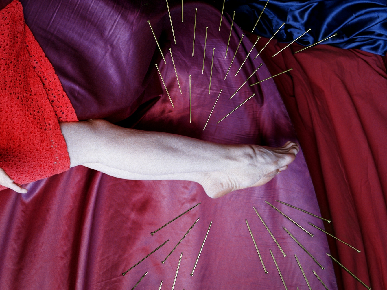 Detail from a larger artwork created with a colour photographic print of a female figure in a bright red dress, set against a purple and blue draped silk background. She is lying horizontal in the frame. Her body is surrounded by groups of dress pins, laid on top of the photographic print. One group attacks her feet and ankles from above and below.