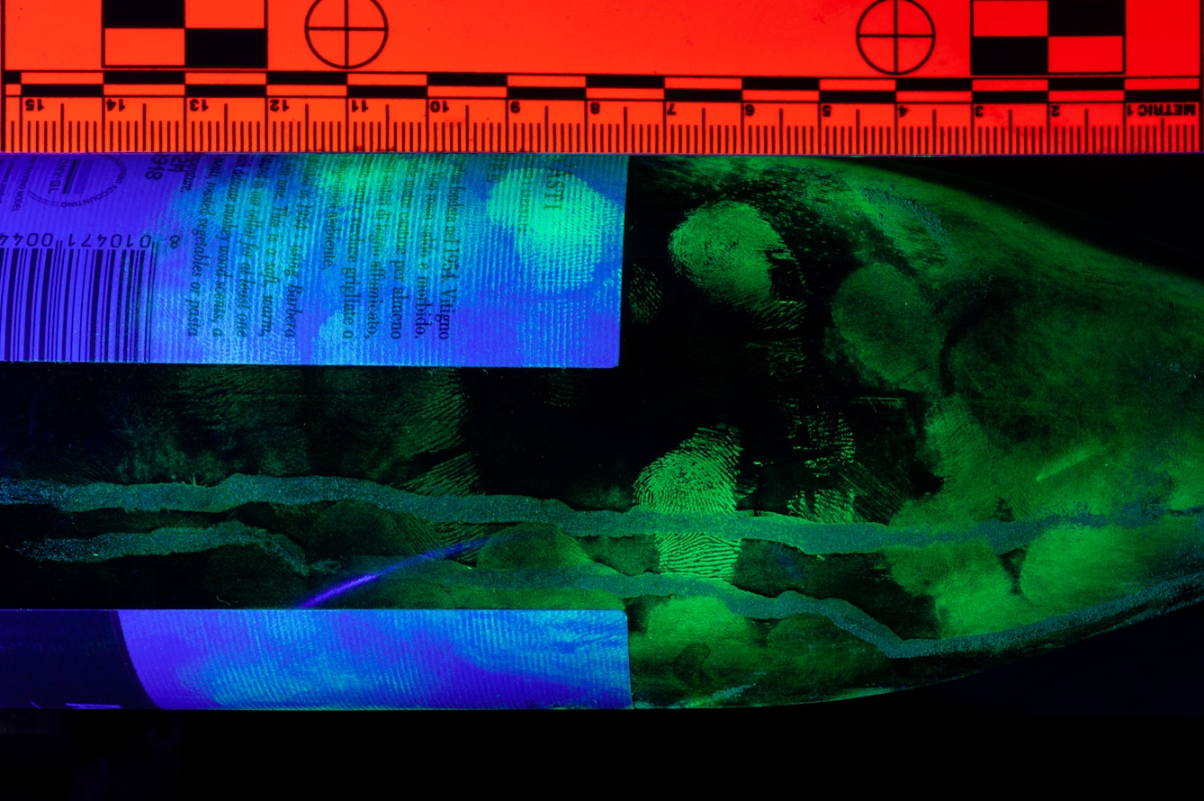 Photograph of part of a wine bottle shot under UV lighting which has reviewed finger prints. Due to the UV lighting the colours in the image are predominantly green and blue. A scale rule in the image has turned bright red.