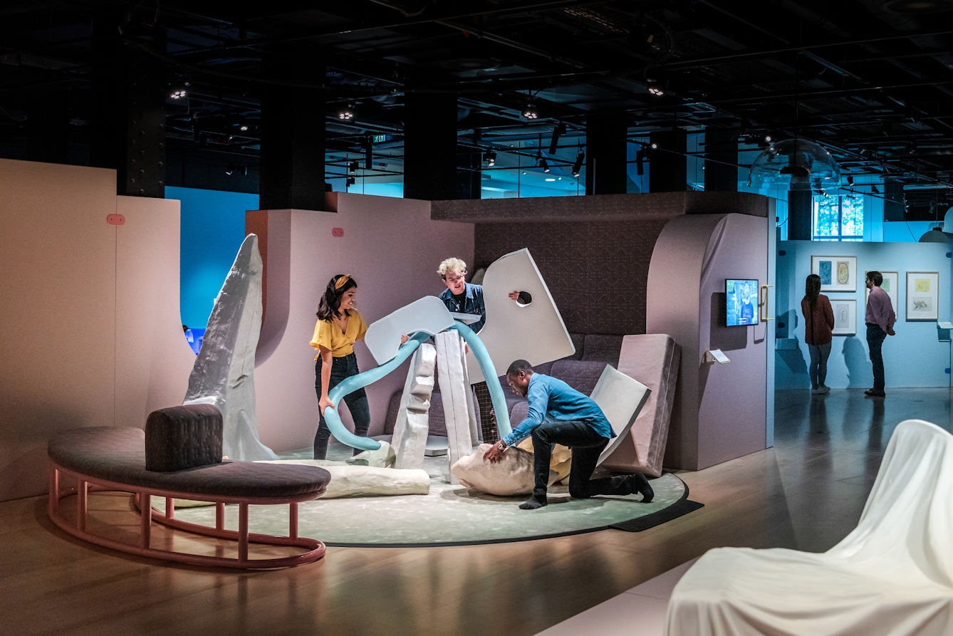 Photograph of an exhibition space showing 3 people working together with large irregular shaped objects to create a sculpture. In the background two other people are exploring other areas of the exhibition.