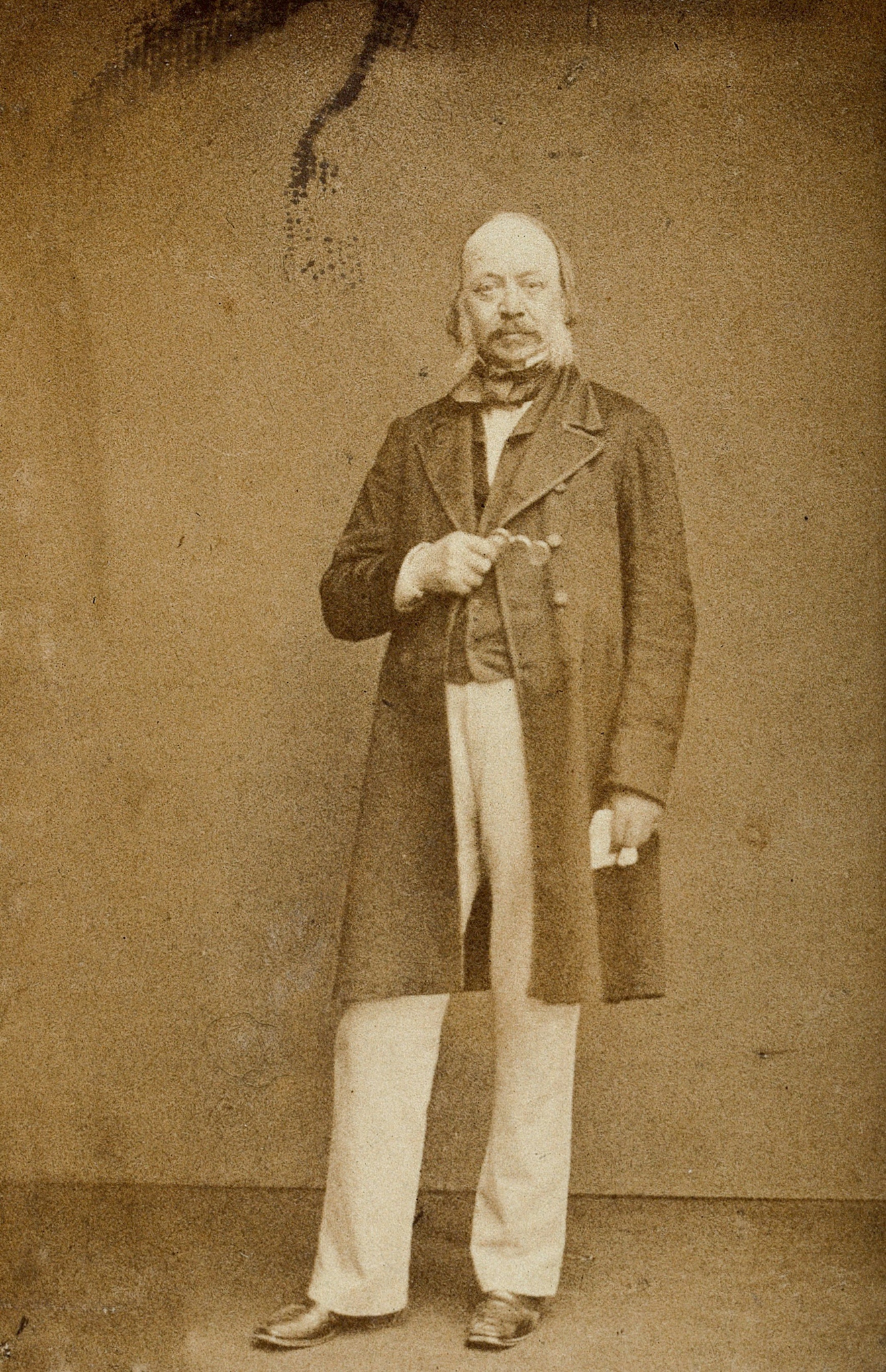 A sepia toned photograph of Sir Edwin Chadwick. A Victorian man with facial hair wearing a long coat with pale trousers and holding pair of glasses.