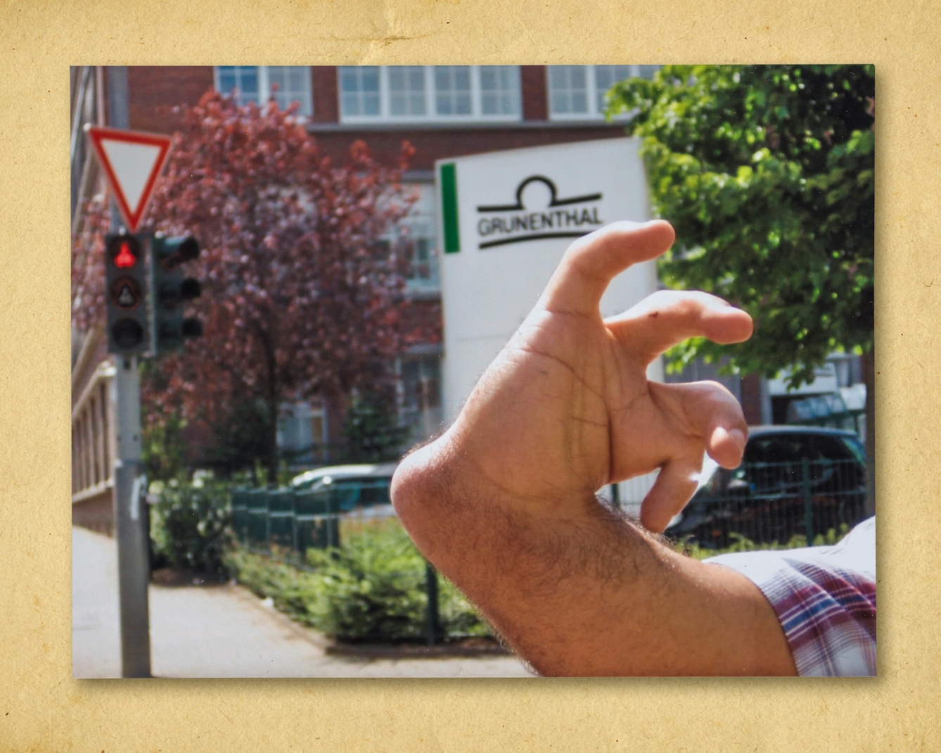 Photograph of a colour photographic print, resting on a brown paper textured background. The print shows a close-up of a man's hand and forearm. His arm, hand and fingers are shaped differently from what might be considered normal. This is as a result of his mother being prescribed thalidomide when she was pregnant. Behind the man's hand in the background is an urban street scene with trees, a set of traffic lights, a traffic sign and a building. In front of the building is a large company sign with the name 'Grunenthal' written within a graphic logo.