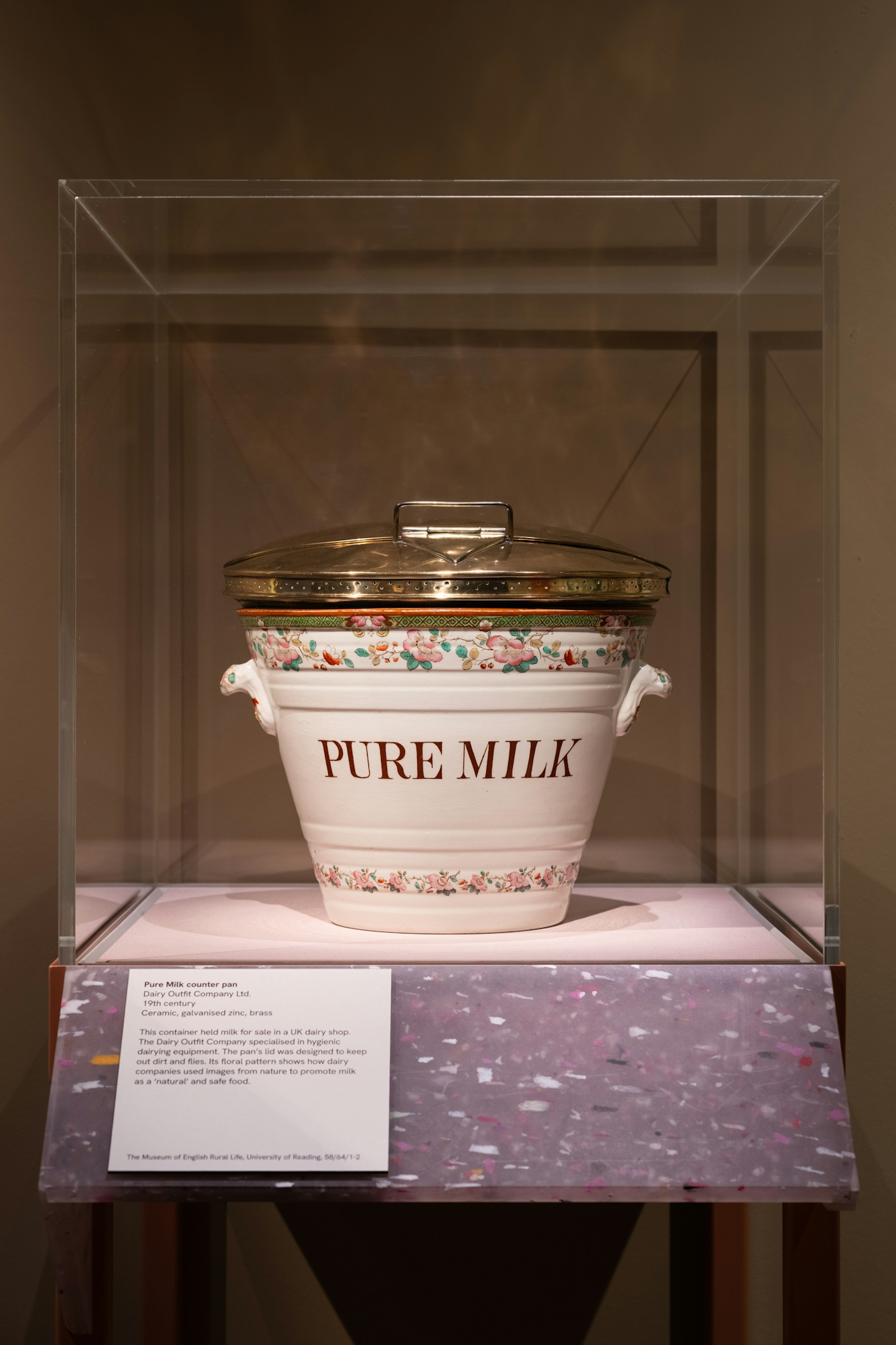 Photograph of a large ceramic milk counter pan with handles and metallic lid. 'Pure Milk' is written on the side of the pan. The pan is displayed in a perspex display case with a caption panel set on a recycled plastic plinth.