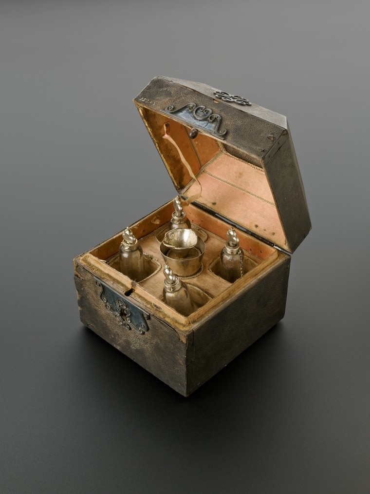 Photograph of small old case, opened to reveal five perfume bottles inside. Ornate clasp.