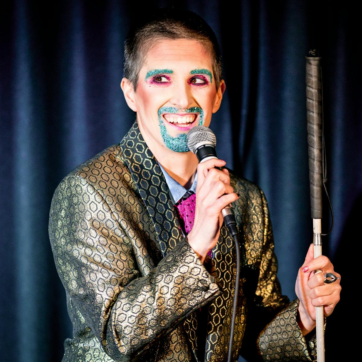 Photograph showing a performer on a stage in a small theatre. The performer is dressed in a shiny gold suit. Their right hand is holding a microphone to their mouth. In their left hand they are holding a white cane. Their mouth and eye brows are covered in blue glitter make-up