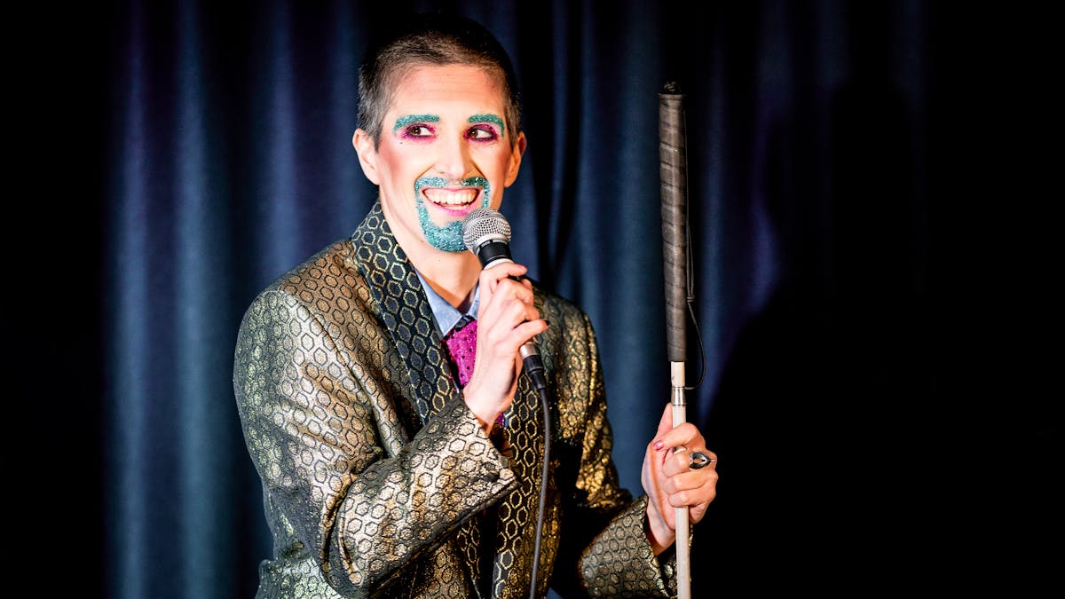 Photograph showing a performer on a stage in a small theatre. The performer is dressed in a shiny gold suit. Their right hand is holding a microphone to their mouth. In their left hand they are holding a white cane. Their mouth and eye brows are covered in blue glitter make-up