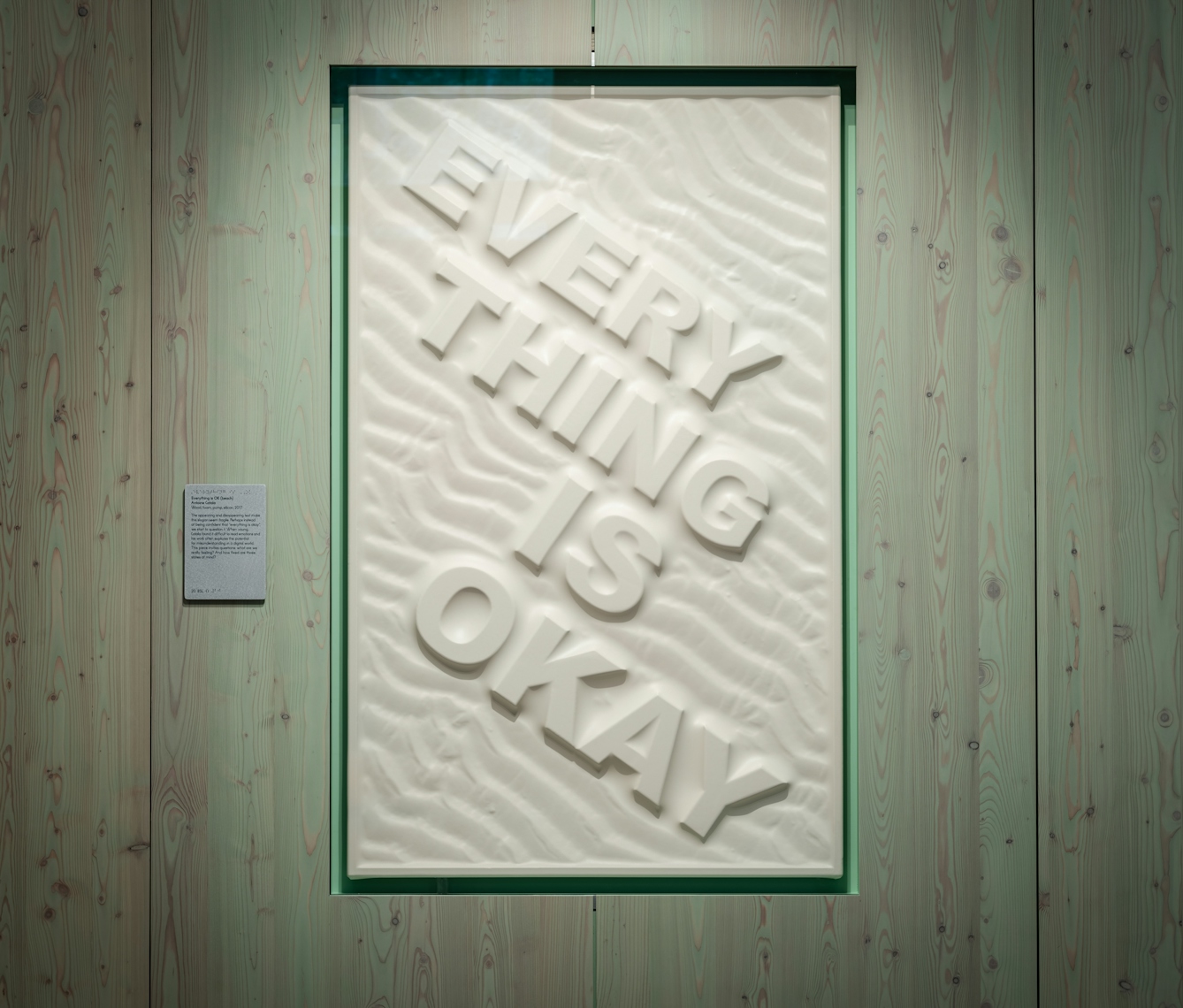A photograph of a large off-white rectangular panel with EVERY THING IS OKAY in relief in large capital letters diagonally across the panel from left to right with one word on each diagonal line. Repeating undulating patterns also move across the panel in the same direction, and the text appears as though a relief in sand. The panel is hung on a light green wood panelled wall.