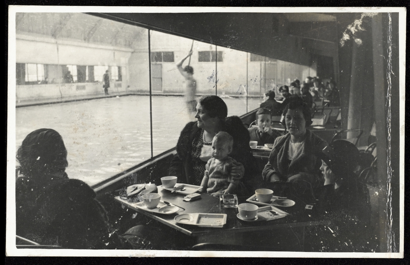 Reproduction of a black-and-white photograph showing a group of women and children at a table having tea watching while people are in the swimming pool at the Pioneer Health Centre