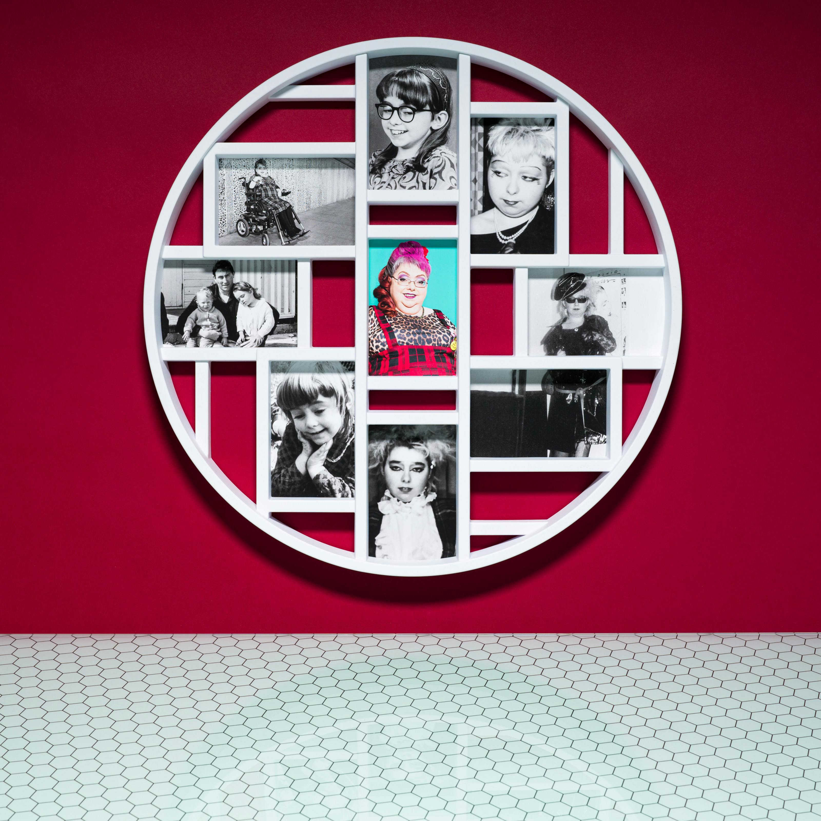 Photograph of a multiframe photo frame containing nine photographs, one in colour and eight in black and white. The frame is hung on a crimson wall above a glass patterned tabletop.