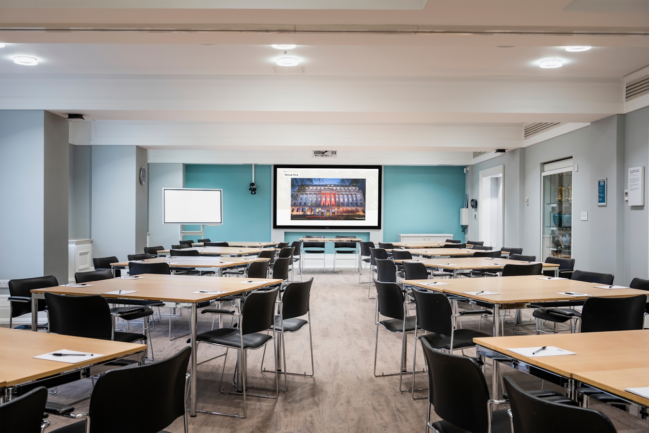 Photograph of the Franks and Steel room at the Wellcome Collection. 

Photograph shows a cabaret set-up, with desks placed in small groups and a whiteboard and presenting screen at the front of the room. 