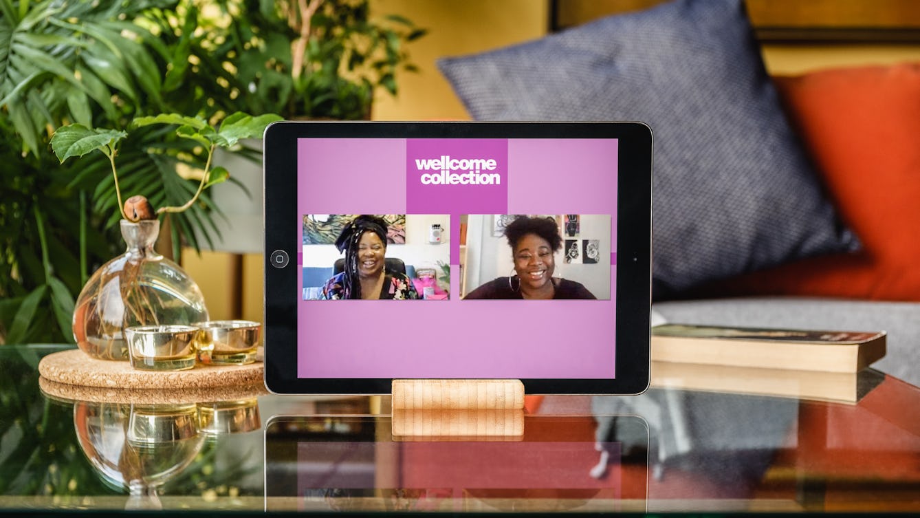 Photograph of a tablet in a stand on a glass coffee table with plants and a sofa behind it. On the tablet screen is a live online event of Cheryl Martin and Rianna Walcott in conversation. The words Wellcome Collection are at the top centre of the screen.