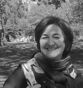 Black and white photograph of a woman with short brown hair looking directly at the camera and smiling. The background is a lawn and trees.