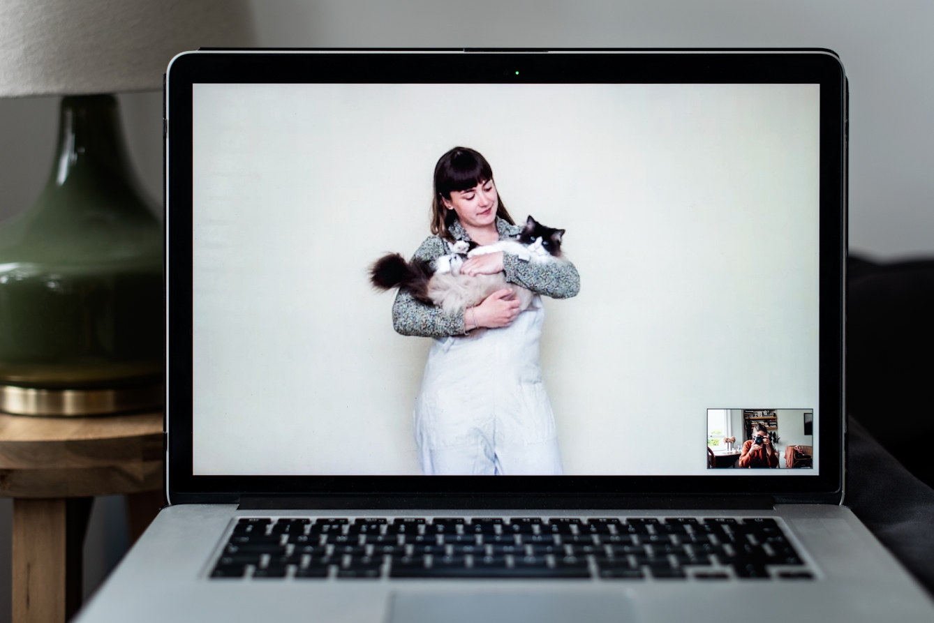 Photograph of an open laptop within a domestic scene. Most of the image is taken up with the screen, with part of the keyboard and trackpad visible. On the screen is a video call showing a woman standing against a white wall looking down at a cat she is holding in her arms. In the bottom right corner of the screen the photographer can be seen in a small floating window, camera to her eye, in the process of taking the picture.