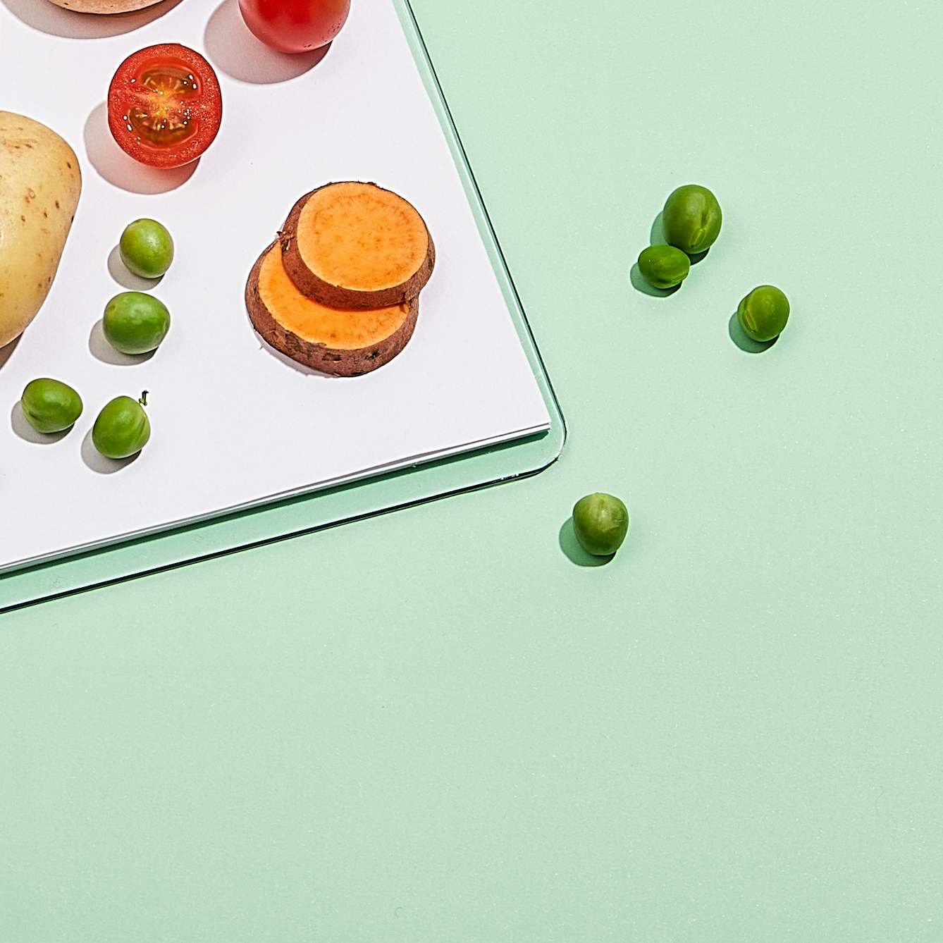 Photograph of the lower corner of a clipboard resting on a light green background. On top of the paper  attached to clipboard is a whole potato, a stack of sliced sweet potato, whole and halved cherry tomatoes and green peas. Loose on the background, to the right of the clipboard are a scattering of loose green peas.