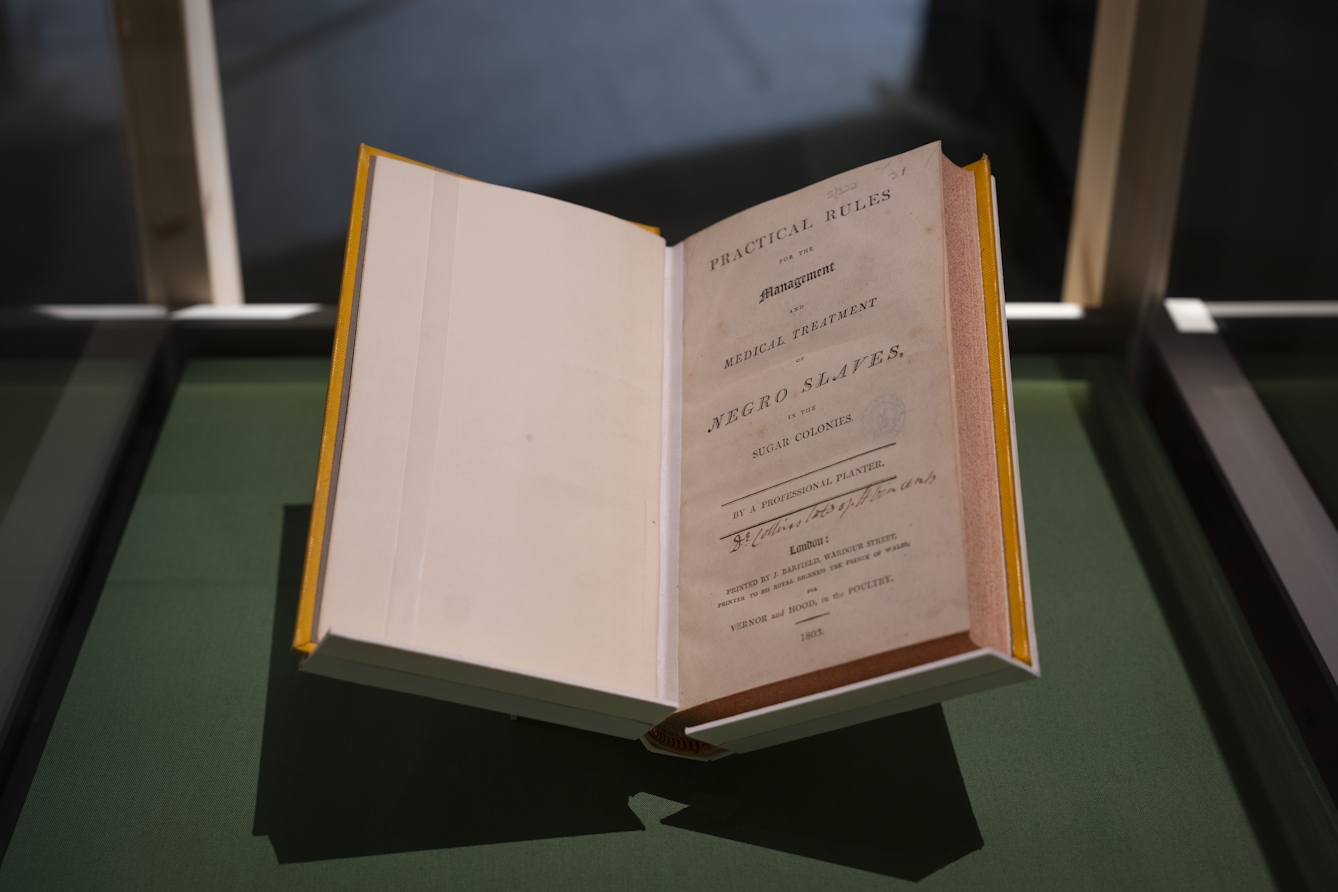A book called 'Practical Rules for the Management and Medical Treatment of Negro Slaves' is laid opened, showing its title page. This opening page says the book was written "by a professional planter", and was printed in London in 1803. It is displayed in a glass case on a green surface. It is a thick book of around 500 pages.
