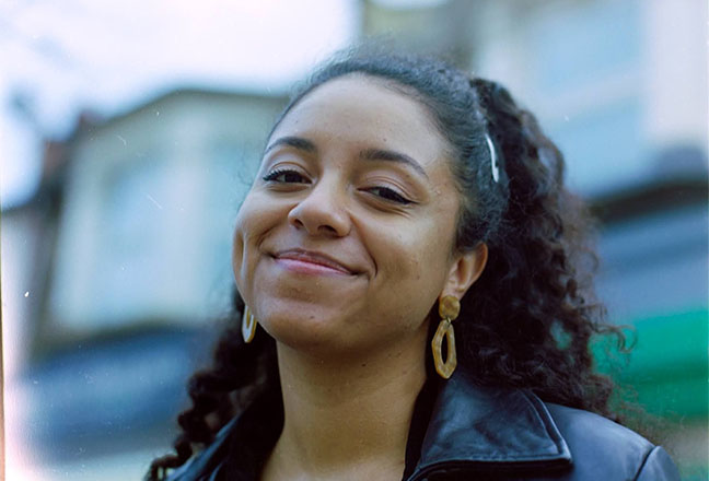 A headshot of Micha wearing gold earrings and a leather jacket, smiling into the camera. 