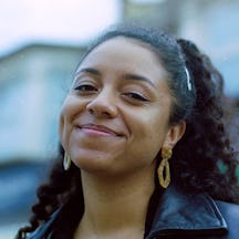 A headshot of Micha wearing gold earrings and a leather jacket, smiling into the camera.
