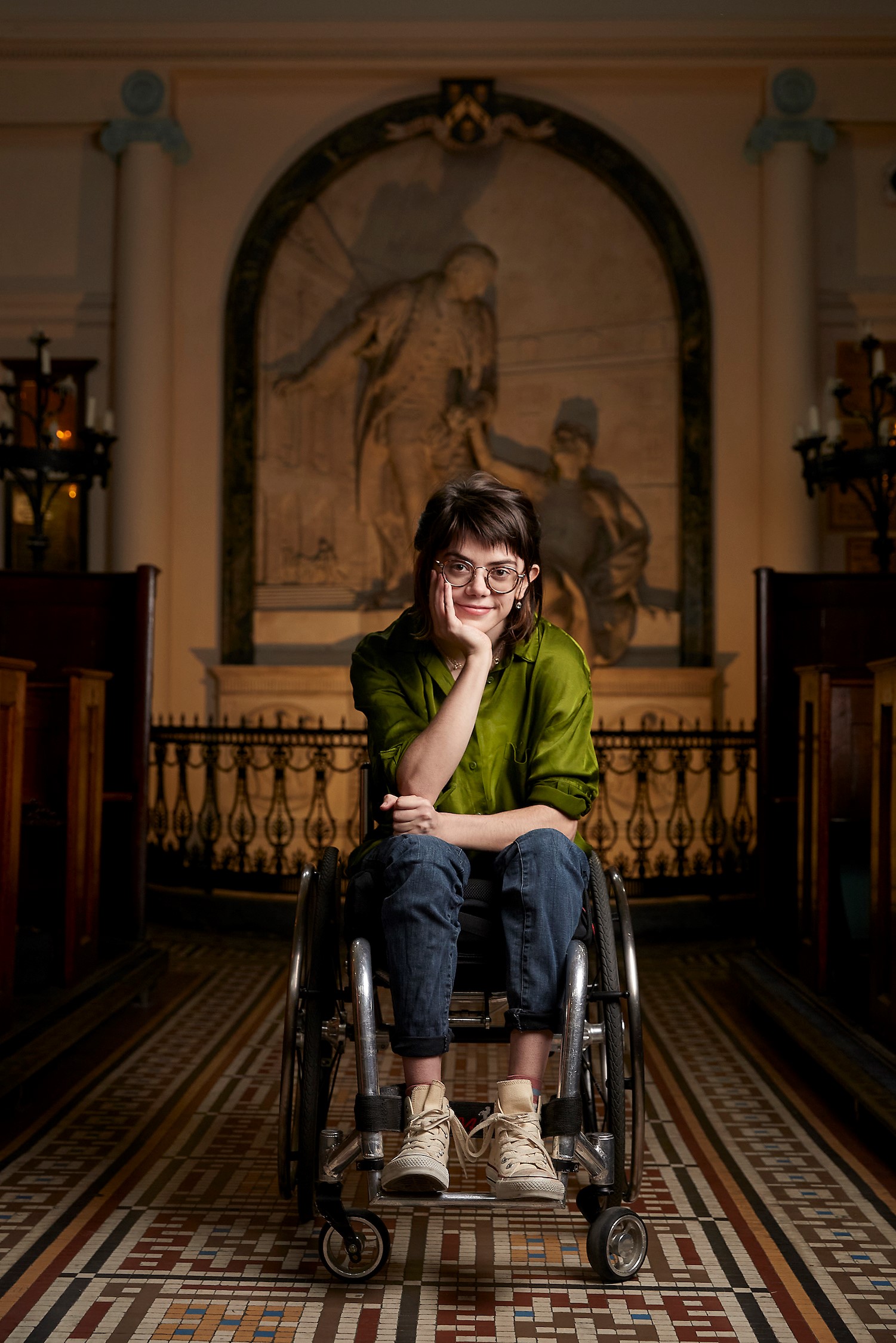 Grace is seated in a wheelchair positioned on a patterned tiled floor inside a grand building. They wear a vibrant green shirt, denim jeans and trainers, and have short brown hair.