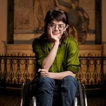 Grace is seated in a wheelchair positioned on a patterned tiled floor inside a grand building. They wear a vibrant green shirt, denim jeans and trainers, and have short brown hair.