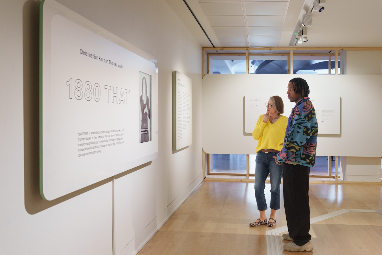 A photograph of two people looking at exhibition information boards and BSL screens.