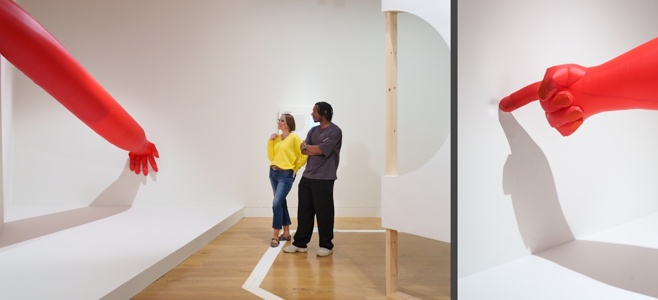 A photograph showing a pair of visitors standing next to an installation of a large inflateable arm, next to this is close-up of an inflatable finger pointing at an indentation in the wall.