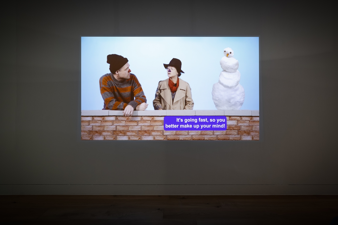 A photograph of a video projection featuring two people with painted noses, gesturing to each other.