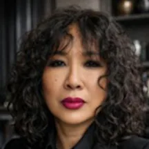 Black and white photo of the head and shoulders of a woman with long dark curly hair. She is looking directly at the camera.