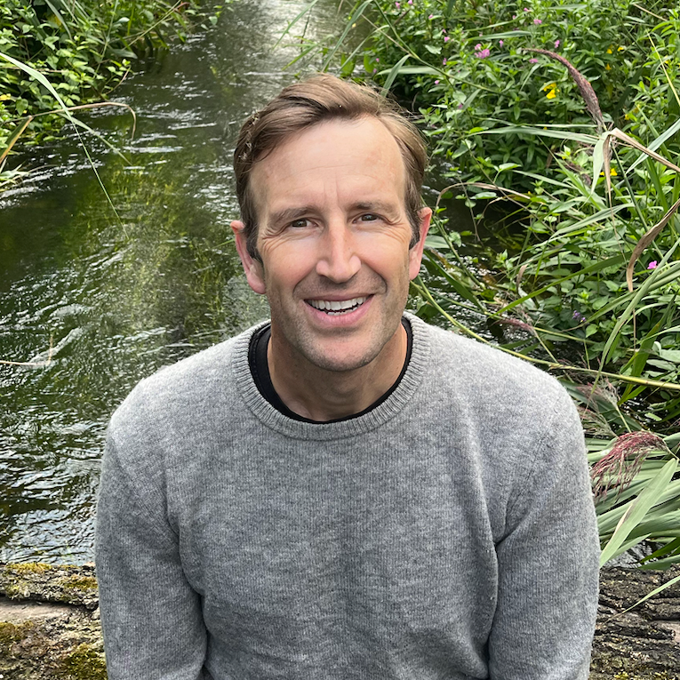 A portrait of Robert Macfarlane looking and smiling directly at the camera. Robert sits in front of a body of water surrounded by green plants.