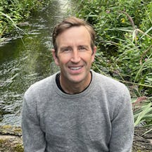 A portrait of Robert Macfarlane looking and smiling directly at the camera. Robert sits in front of a body of water surrounded by green plants.