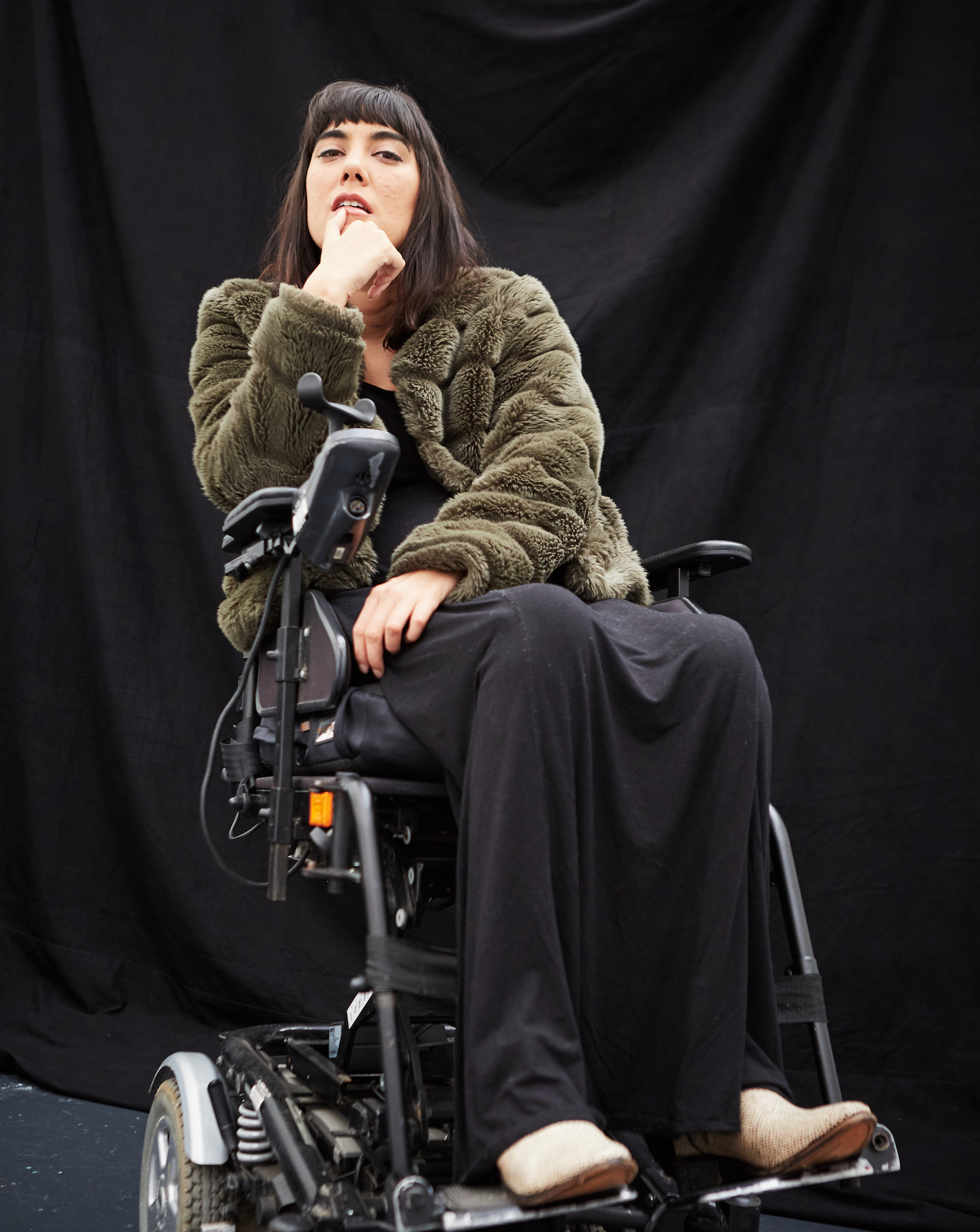 A photograph of Kyla who has short, dark hair and is wearing a green jacket, sitting in a wheelchair. 