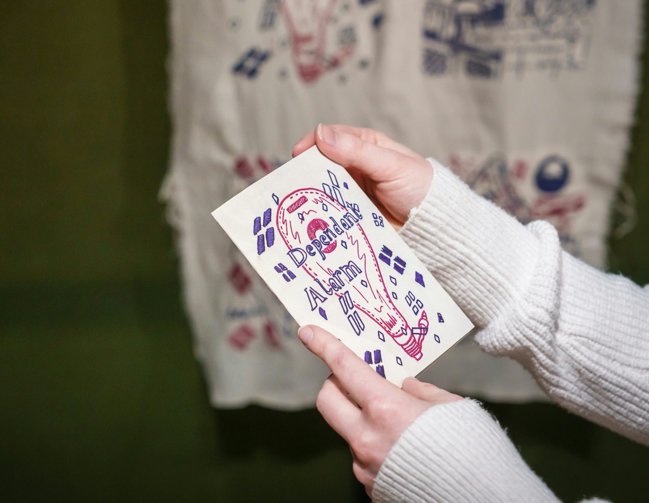 A gallery visitor holds the paper zine titled 'Dependant Alarm' in their hands, in front of an embroidered version of the zine hanging on the gallery wall.