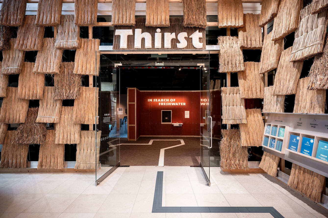 The atrium outside Gallery 1 at Wellcome Collection featuring a thatched wall and large sign above the doorway that reads 'Thirst'.