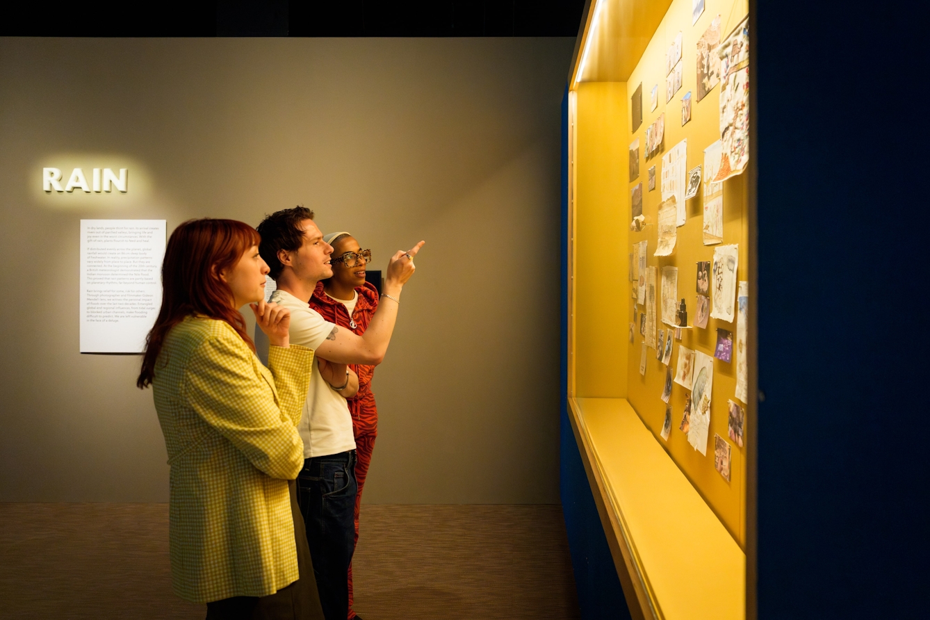 A group of three people standing in front of and observing a wall display of paintings, tapestries / embroideries and photographs.