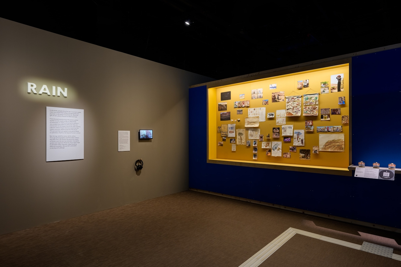A large ceramic sign that reads 'Rain' with a display of paintings, tapestries / embroideries and photographs to the right.