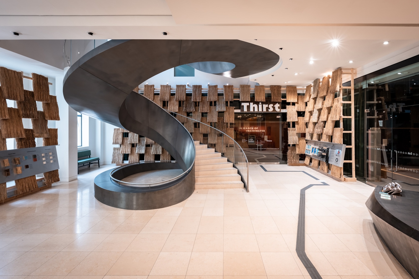 The atrium outside Gallery 1 at Wellcome Collection featuring a thatched wall; a large sign above the doorway that reads 'Thirst' and a spiral staircase.