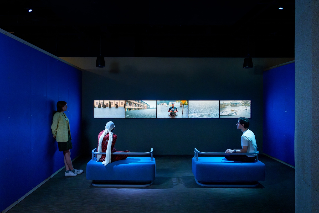 Two people sitting on benches and another leaning against a wall watching a series of videos on five screens.