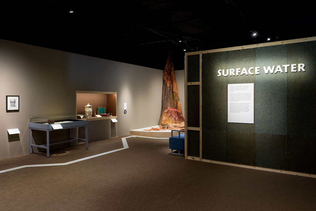 A large ceramic sign on a wall that reads 'Surface Water' with a couple of display cases to the left and a large fabric artwork in the background.
