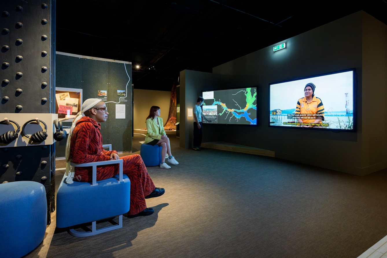 Two people sitting on benches, crossed-legged, watching a film and another interacting with an adjacent screen.