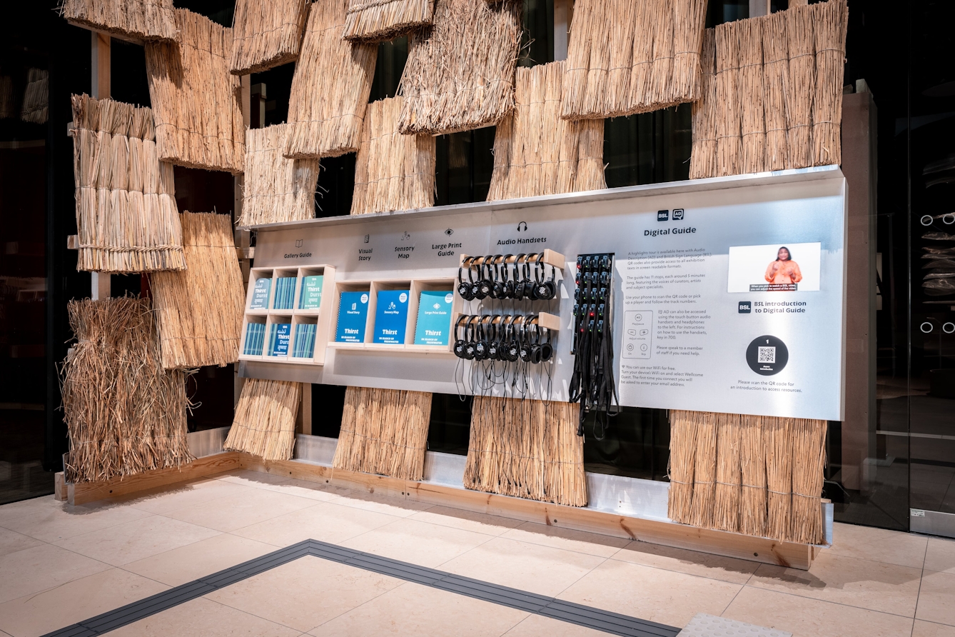 A wall covered in thatch with some shelves storing printed versions of the gallery guide, visual story, sensory map and large print guide for the Thirst exhibition. There are also headphones and handsets.
