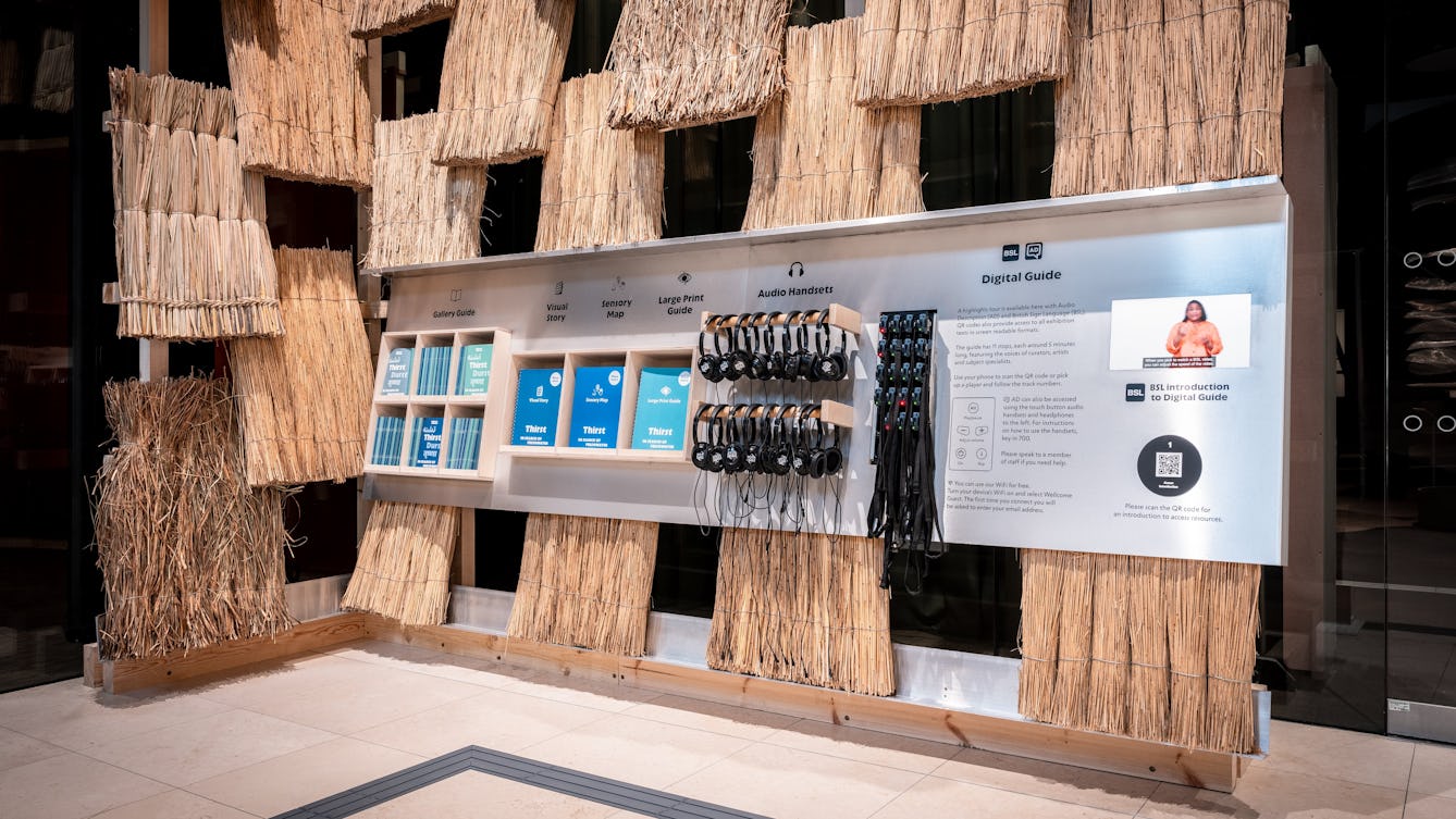 A wall covered in thatch with some shelves storing printed versions of the gallery guide, visual story, sensory map and large print guide for the Thirst exhibition. There are also headphones and handsets.