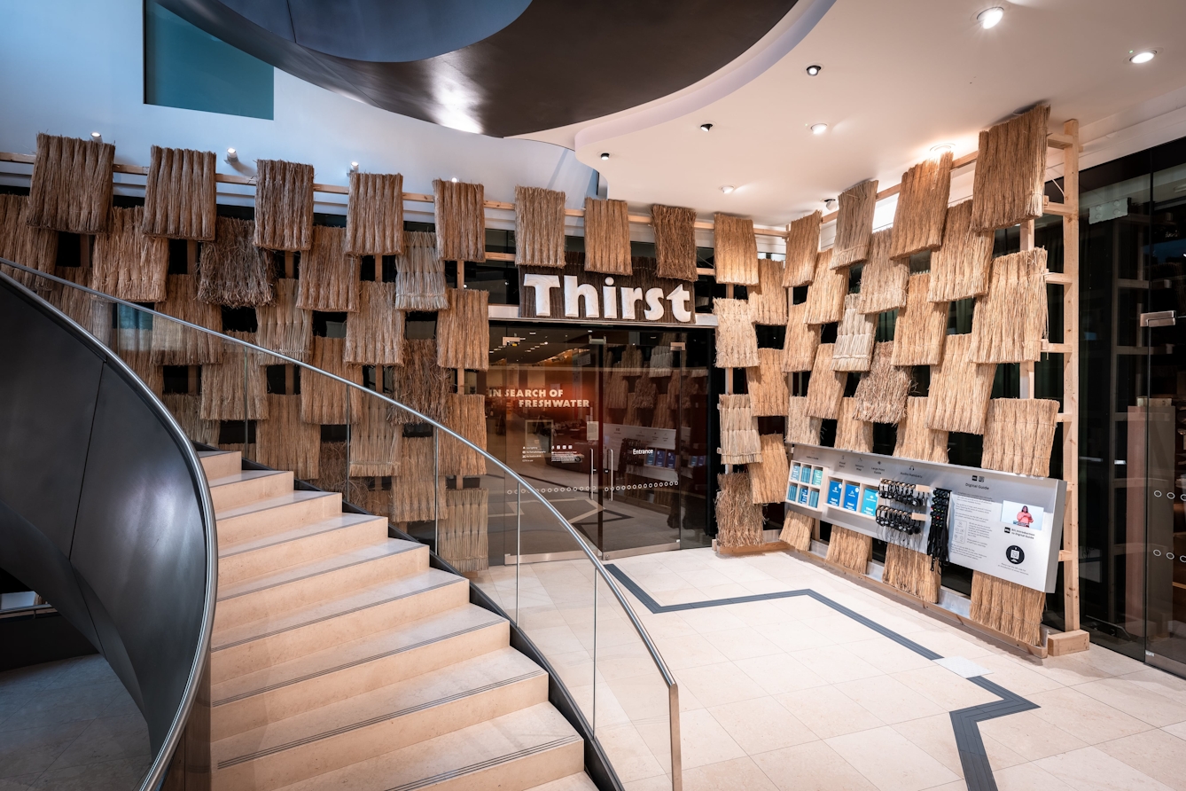 The atrium outside Gallery 1 at Wellcome Collection featuring a thatched wall and large sign above the doorway that reads 'Thirst'.